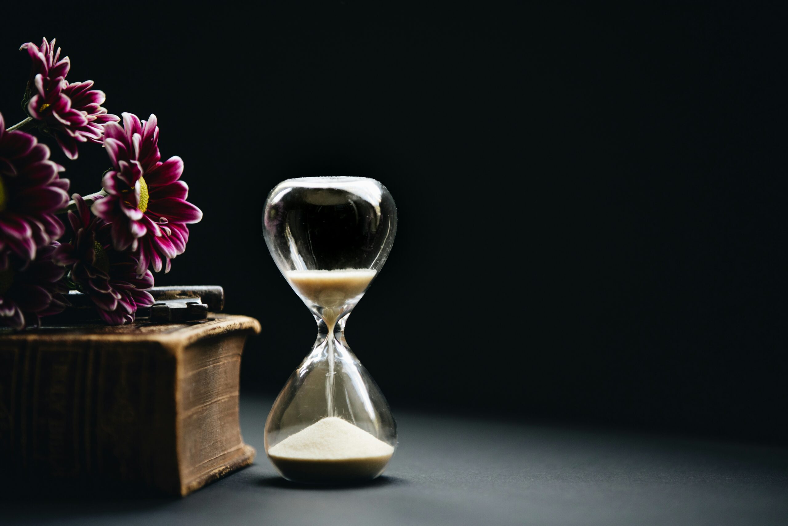 an hourglass next to a book and flowers in front of a dark gray background